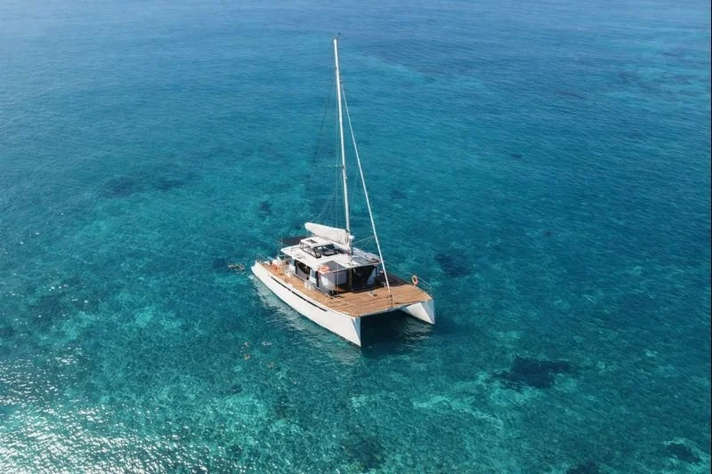 An aerial view of a white sailing catamaran on clear blue ocean water.