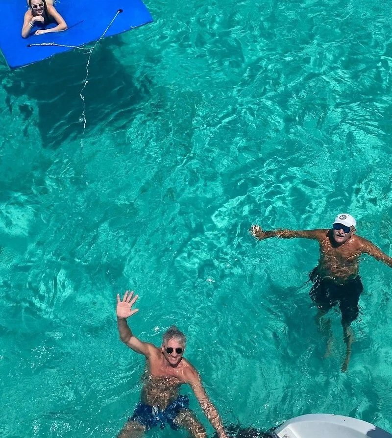 Two men are swimming and waving in clear blue water near a catamaran.