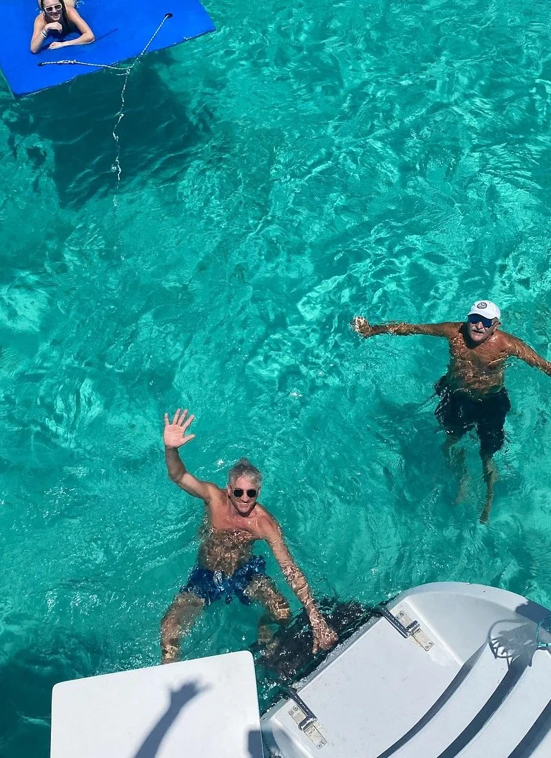 Two men are swimming and waving in clear blue water near a catamaran.