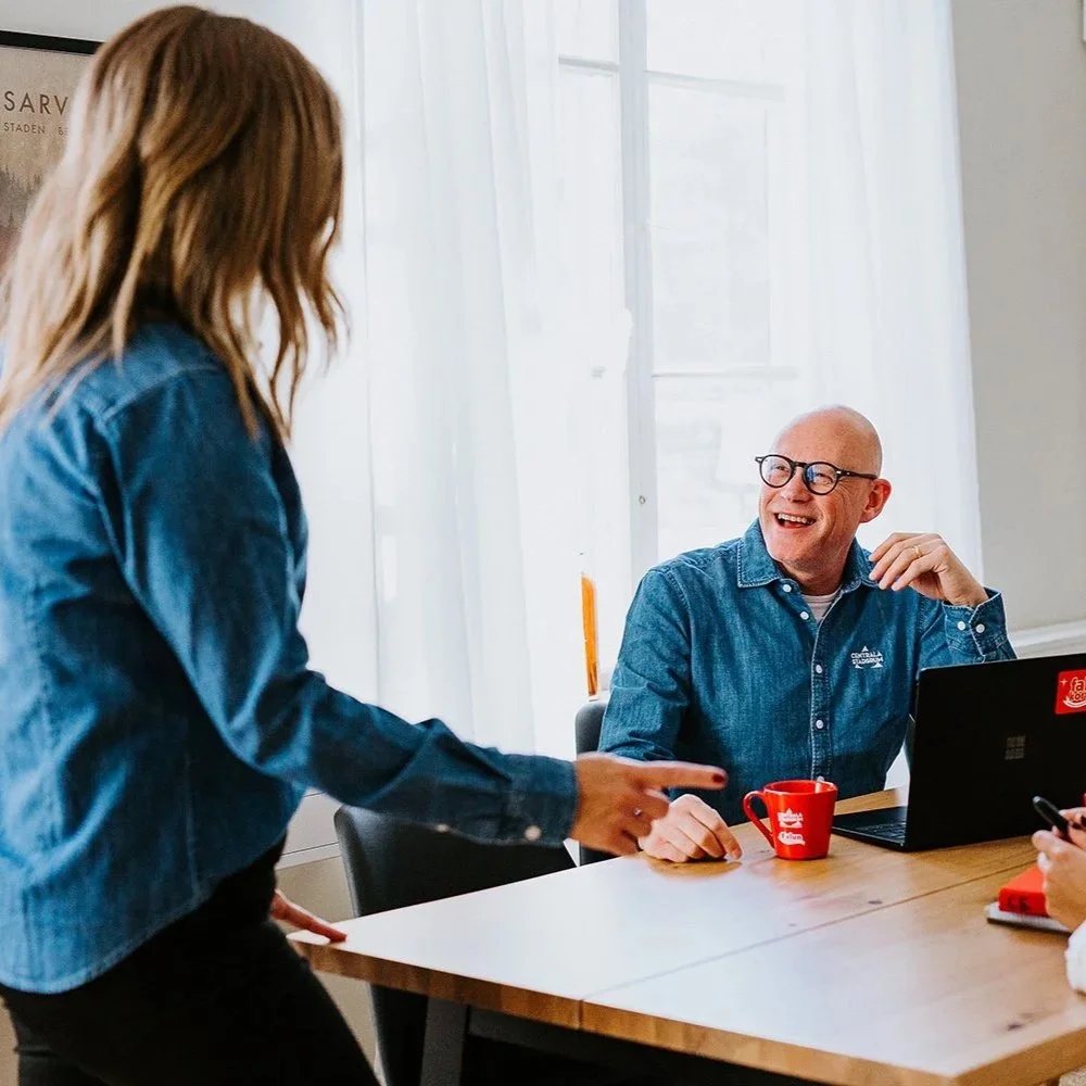 En kvinna på Centrala Stadsrum står och pratar med en man som sitter vid ett bord. Mannen ler och har en röd kopp framför sig, de ser ut att ha ett samtal i ett kontorsmiljö.