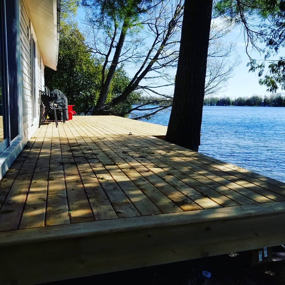 Newly built wooden deck extending from a house by a body of water, with a large tree in the background and a view of the water and distant trees.