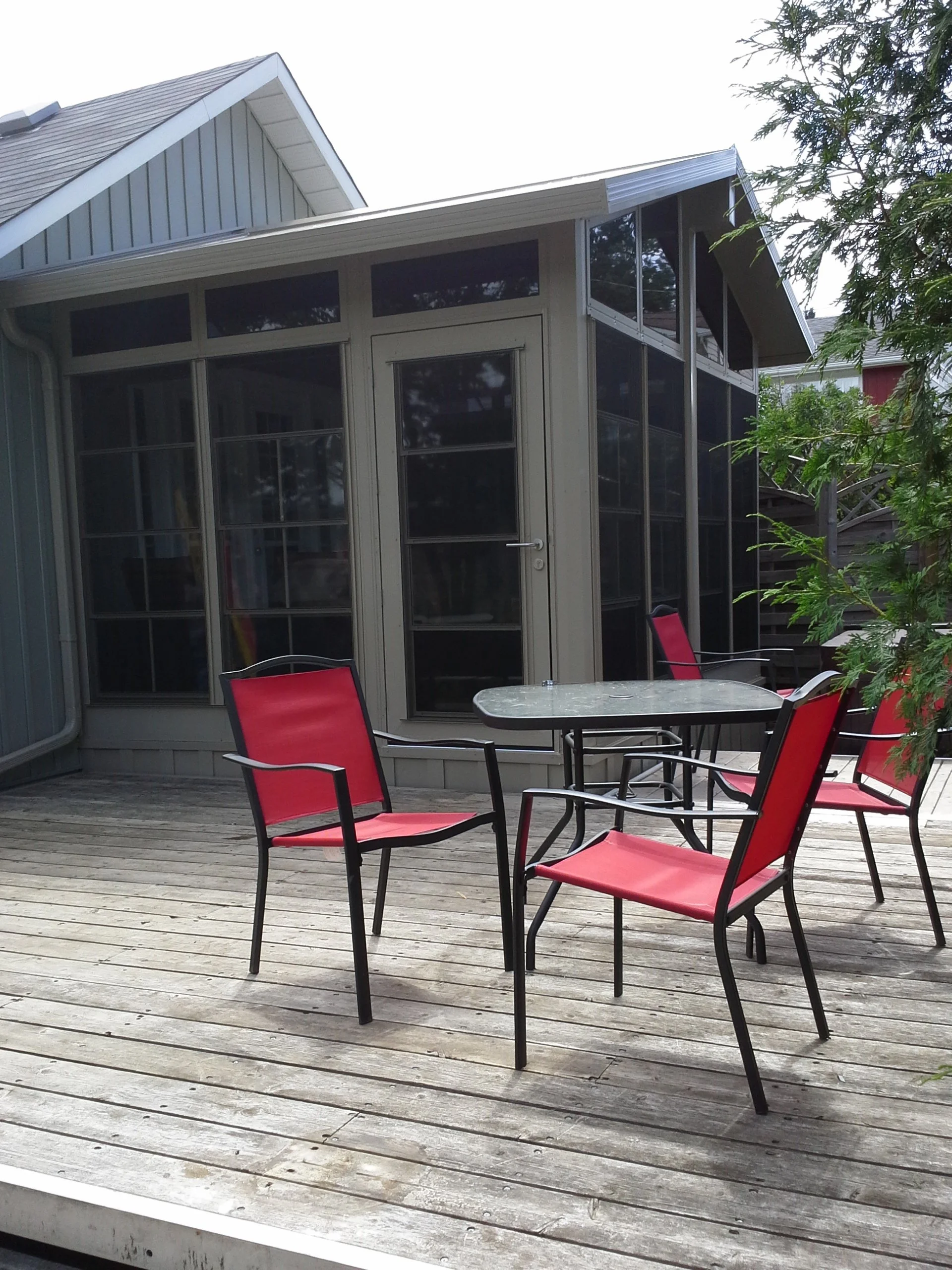 Outdoor patio with a wooden deck, a round table, and four red chairs. A screened porch attached to a house with gray siding and a gabled roof is in the background. There are trees and greenery around.
