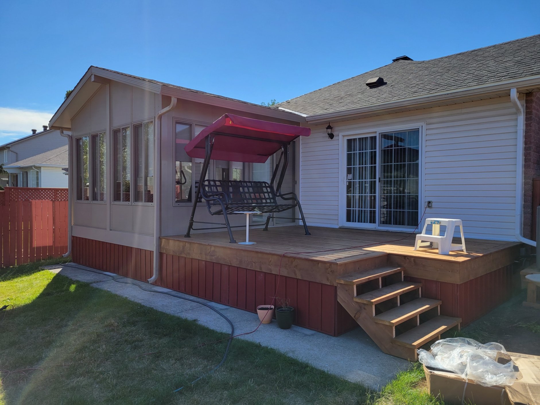 Backyard deck with stairs, a swing with a pink canopy, a small white step stool, and potted plants, attached to a house with white siding and sliding glass door, under a clear blue sky.