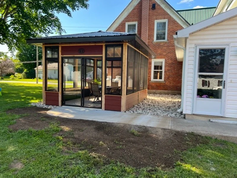 A backyard scene with a screened-in porch attached to a brick house and a small white outbuilding, surrounded by a grassy lawn and trees.
