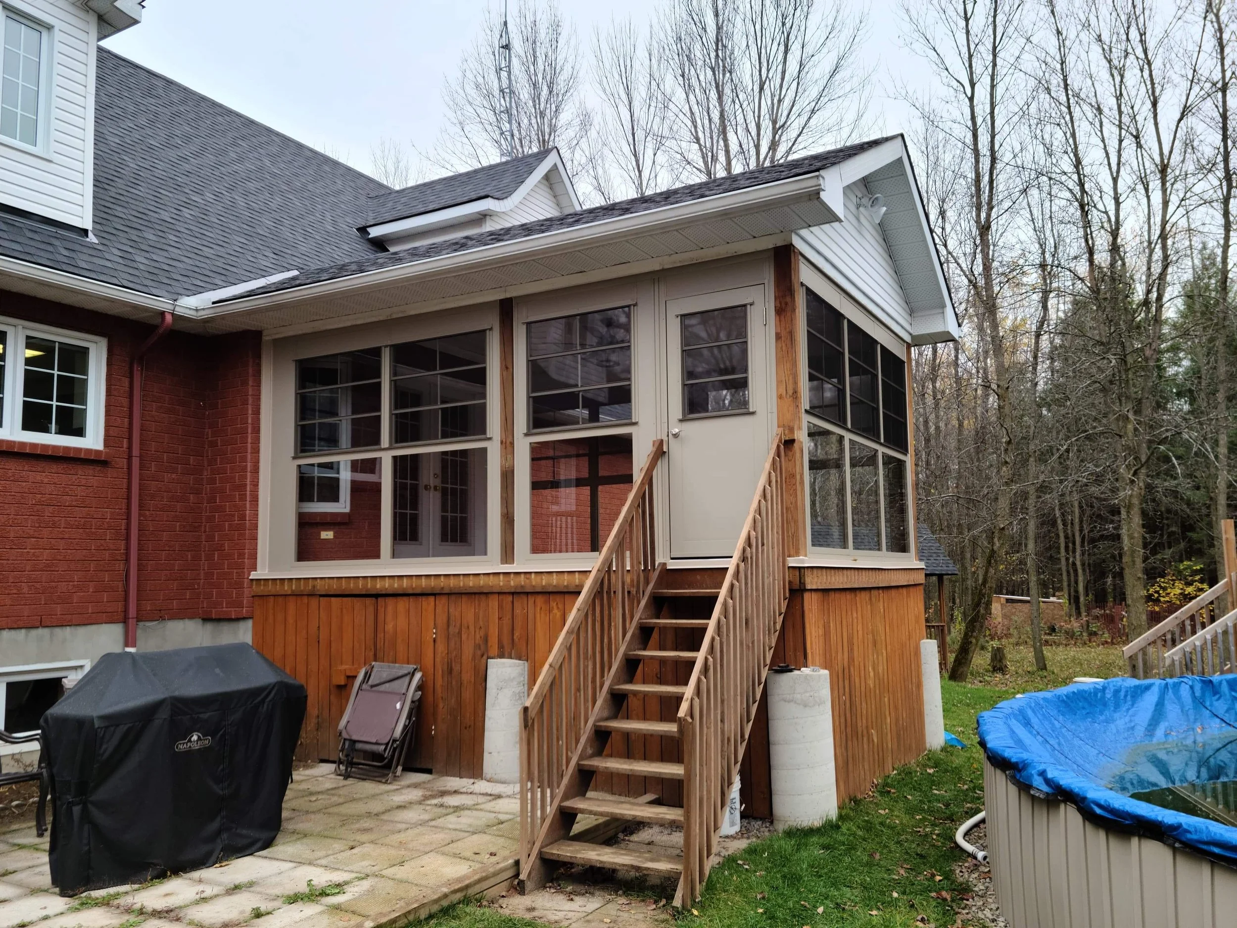 Back patio of a house with a screened porch above a wooden deck. There are outdoor furniture, a grill, and a small swimming pool.