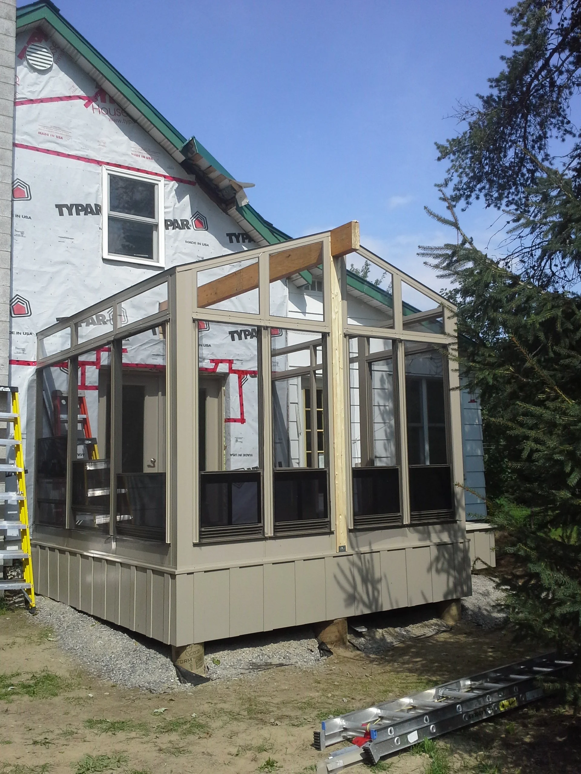 A house under construction with a screened-in porch being built. The house has a skylight window and is covered with house wrap, with a roof partially installed. There are ladders and construction materials nearby.