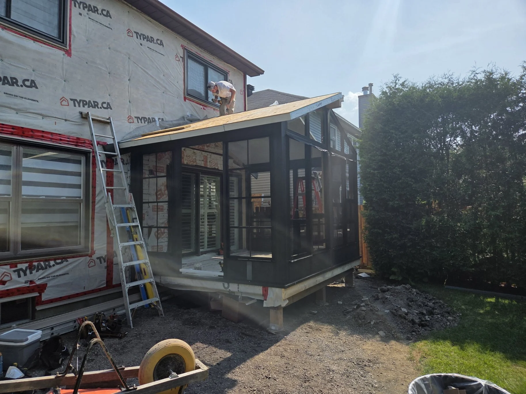 Construction worker installing roofing on a house extension with a screened porch, ladder leaning against the house, and building materials and tools on the ground.