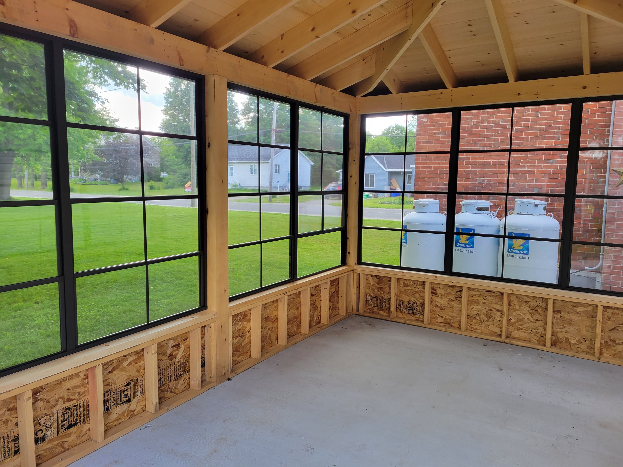 Interior of a porch or sunroom under construction with large windows, showing a green lawn and neighboring houses outside.