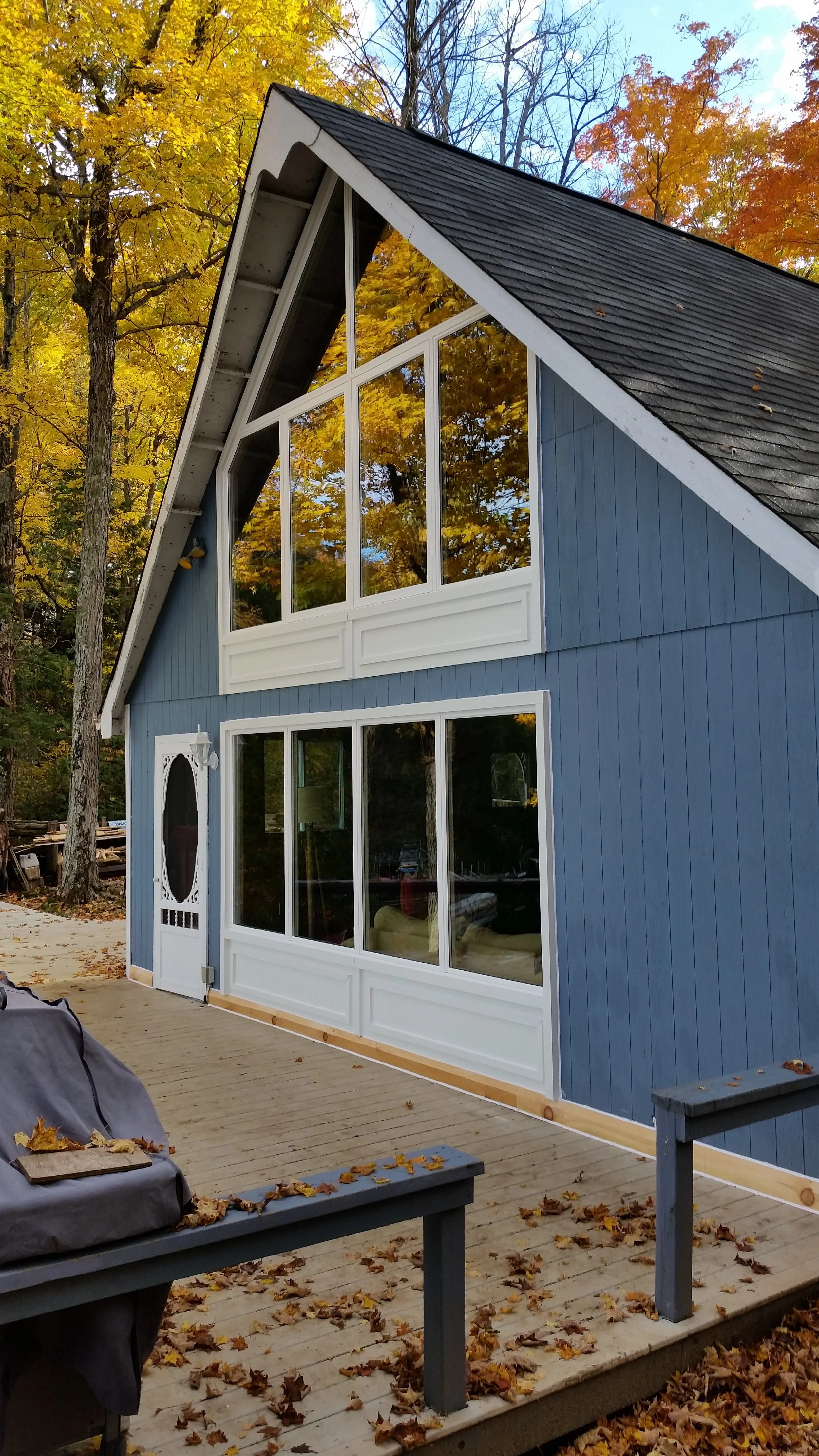 A two-story house with blue vertical siding, large front windows with white trim, and a steeply pitched roof, in a wooded area during autumn with colorful fall foliage.