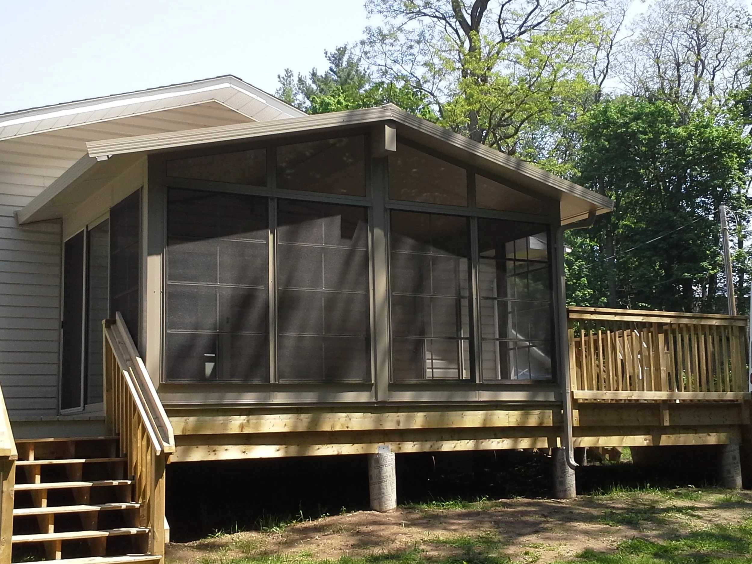A house with a screened-in porch and wooden stairs leading up to it, surrounded by trees and greenery in a backyard.