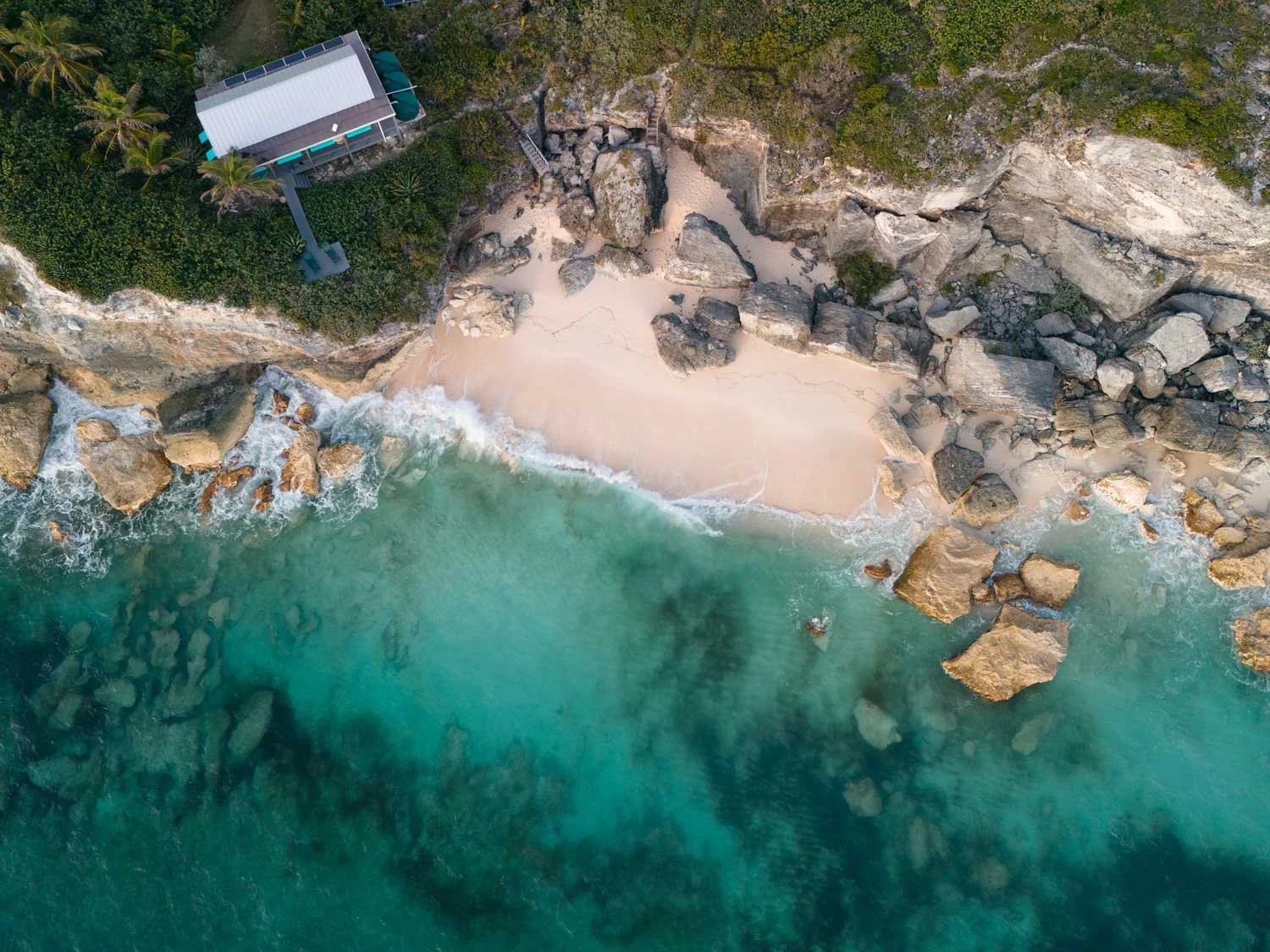 Aerial view of a coastal scene showing a small sandy beach, rocks, turquoise water, a wooden staircase leading to the beach, and a house among trees.