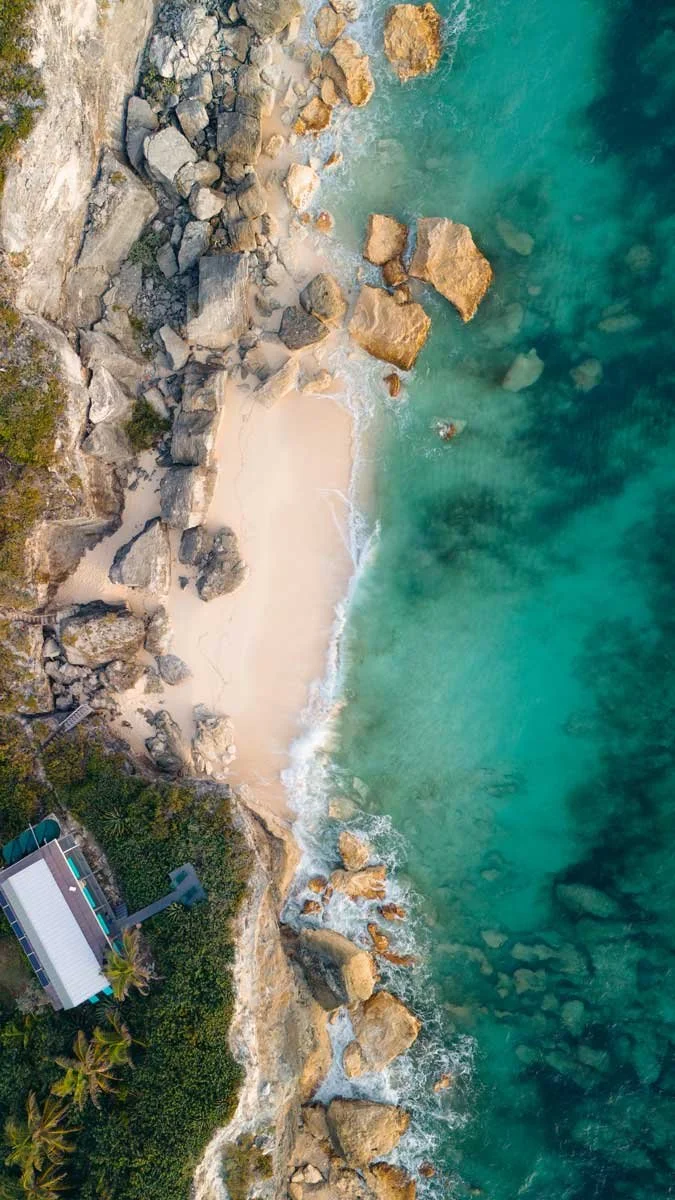 Aerial view of a rocky beach with clear blue-green water and a small sandy area, surrounded by greenery and a building with a pool.