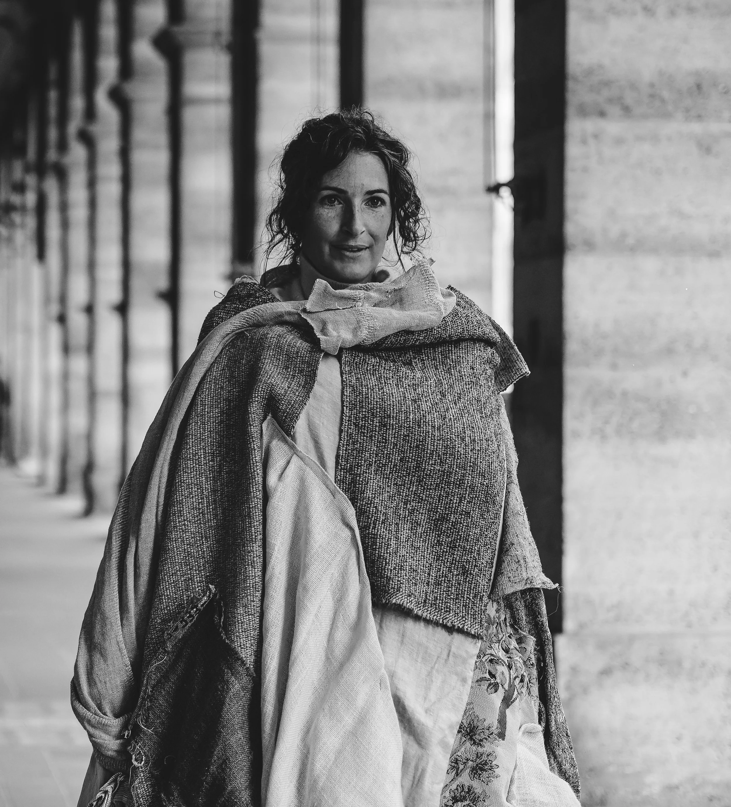 Een zwart-wit foto van een vrouw met donker, krullend haar die naar de camera kijkt. Ze draagt meerdere lagen kleding en staat buiten, met een gebouw op de achtergrond.