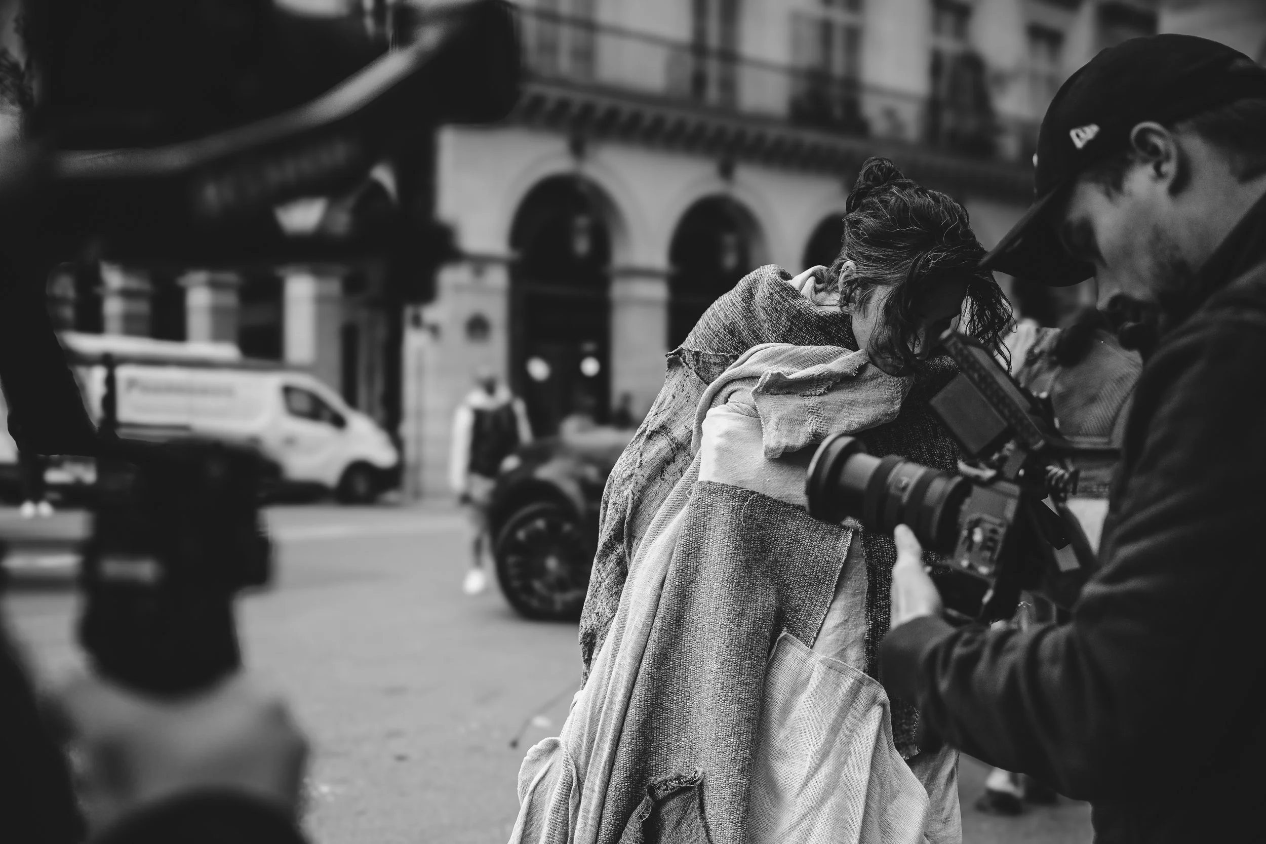 Fotografie van een filmmaker die een vrouw filmt op straat, met oude gebouwen en voertuigen op de achtergrond, zwart-wit.