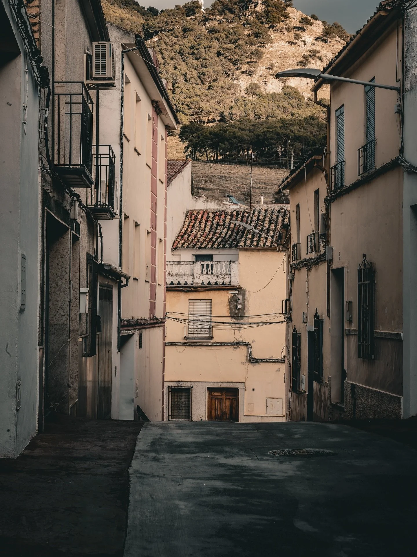 Een smalle straat tussen oude, gekleurde huizen met balkons en hangende kabels, met een heuvel en bomen op de achtergrond.
