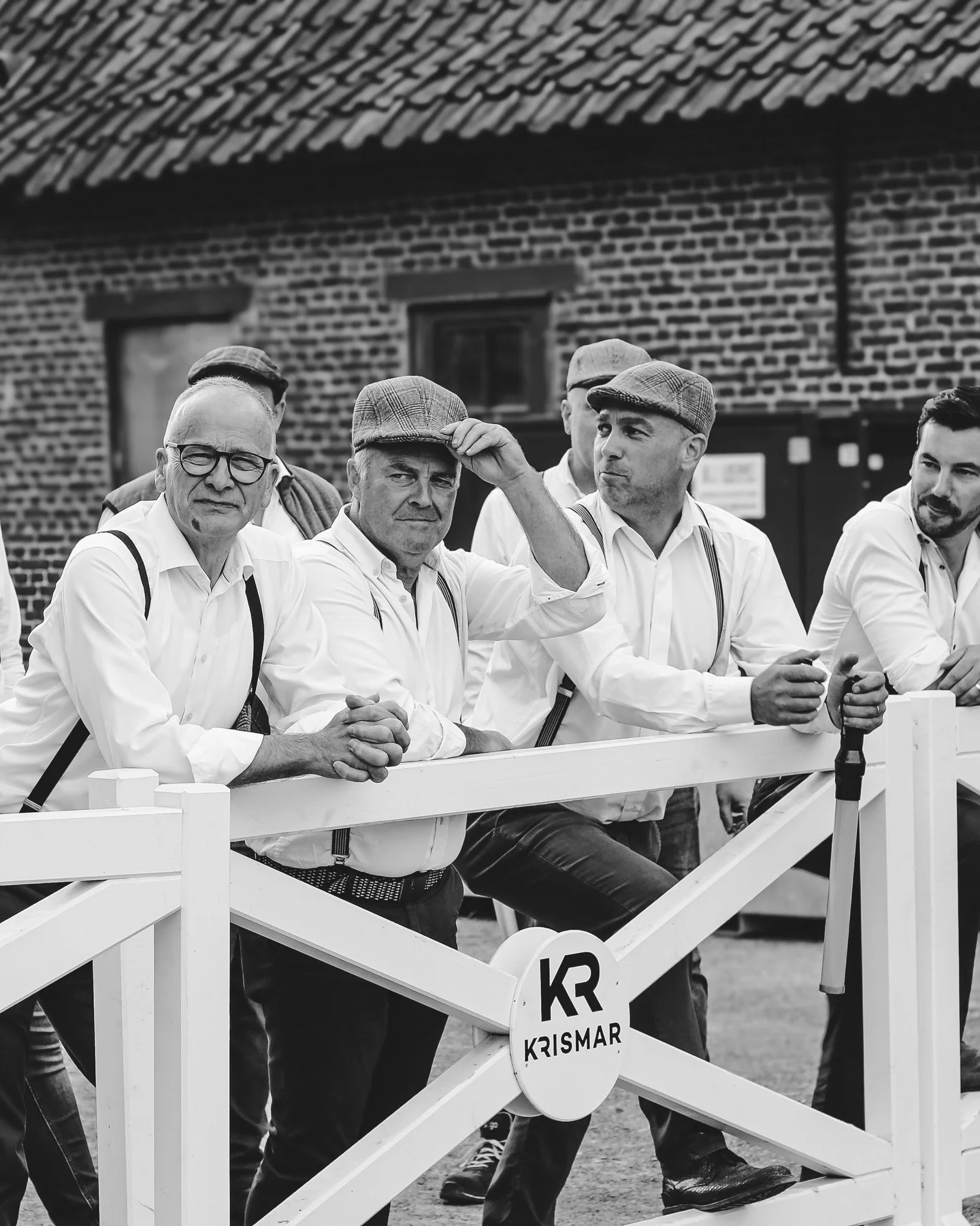 Zes mannen in vintage stijl, gekleed in witte overhemden en flat caps, staan achter een witte schutting met 'KRISMAR' logo. Ze lijken in een dorp of op het platteland, met een bakstenen gebouw en een dak met dakpannen op de achtergrond.