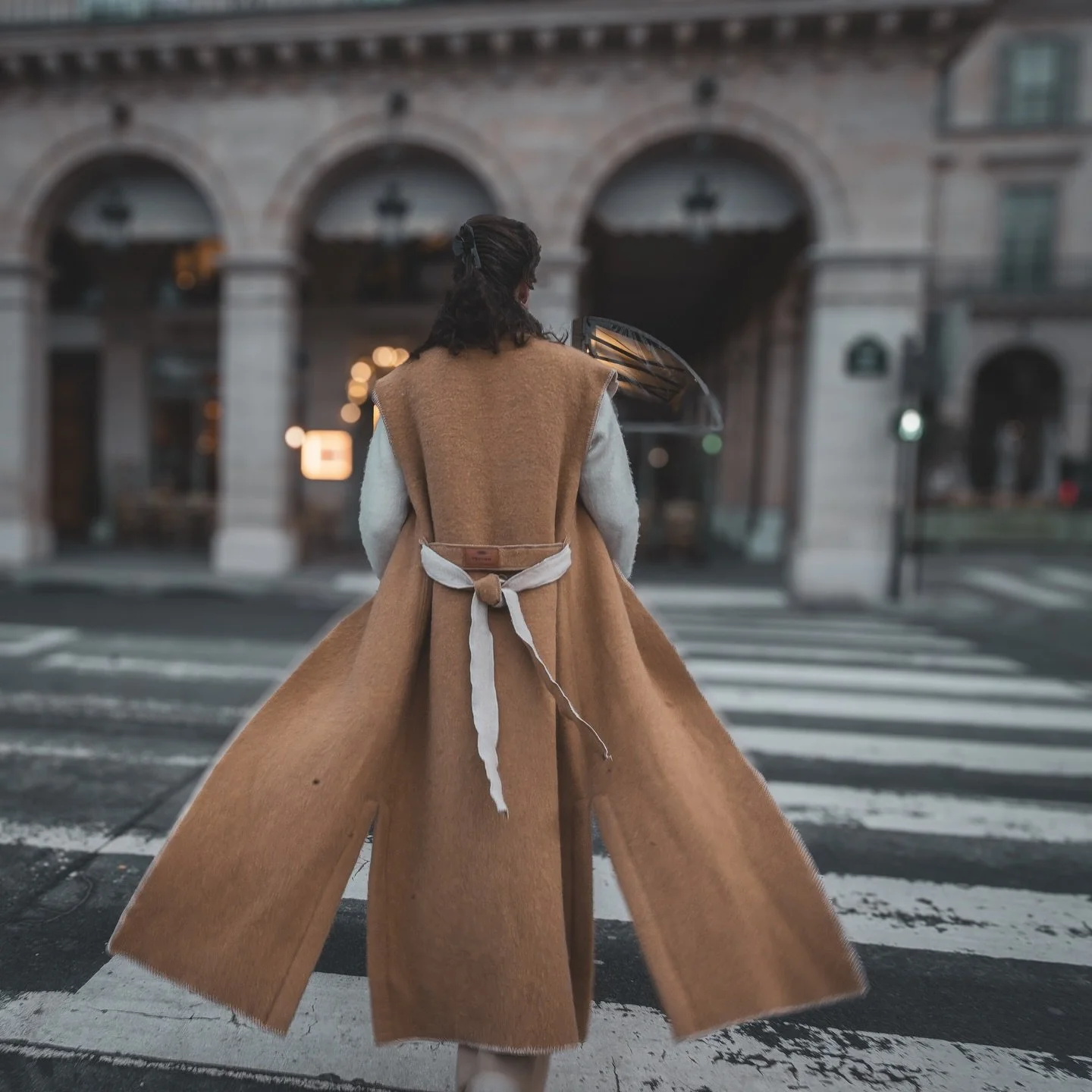 Persoon in een lange, bruine jas met witte mouwen wacht op een zebra overgang op een straat in een stad, met oude gebouwen op de achtergrond.