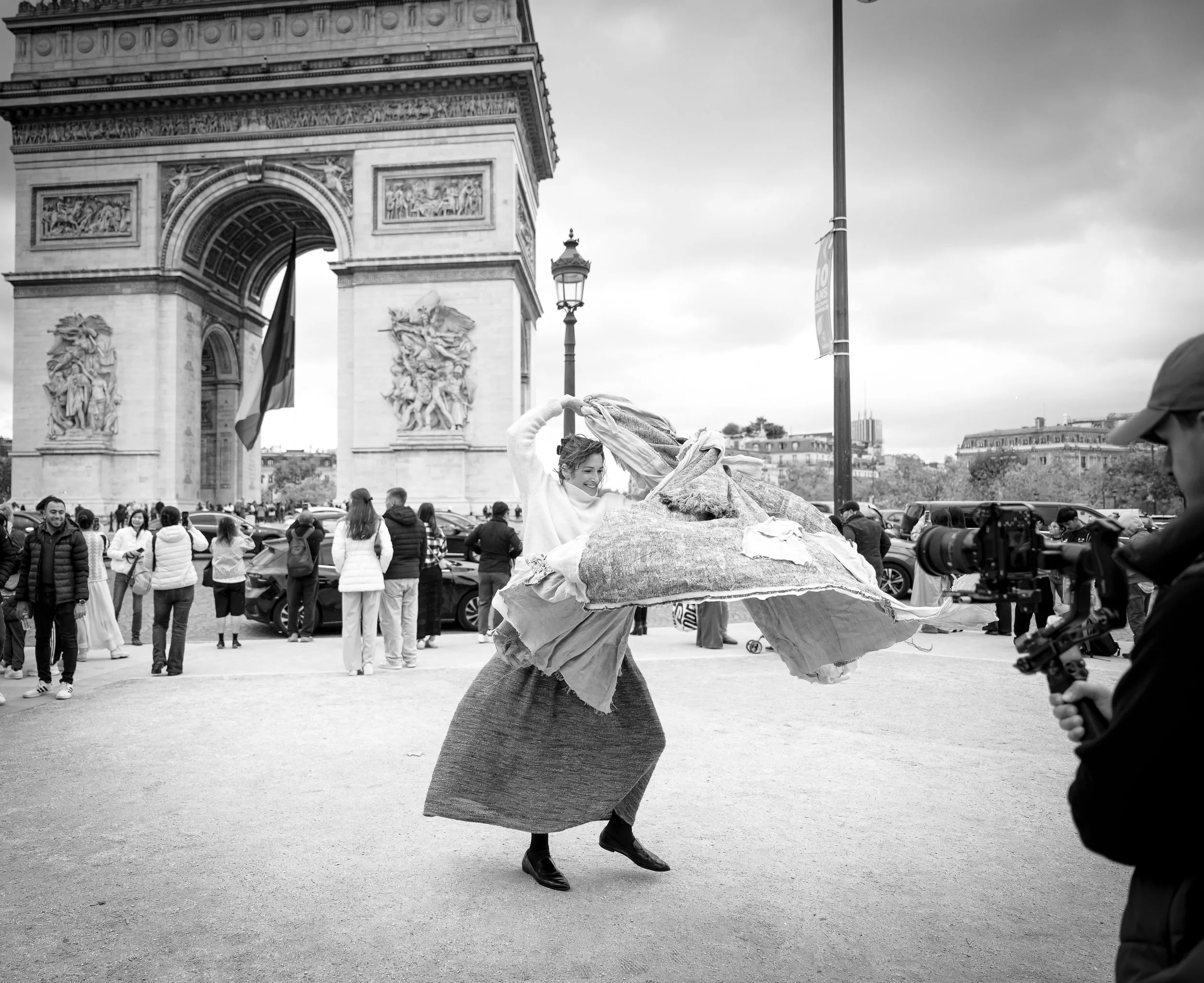 Een vrouw danst vrolijk voor de Arc de Triomphe in Parijs, omringd door een groep mensen en een cameraman, op een bewolkte dag.