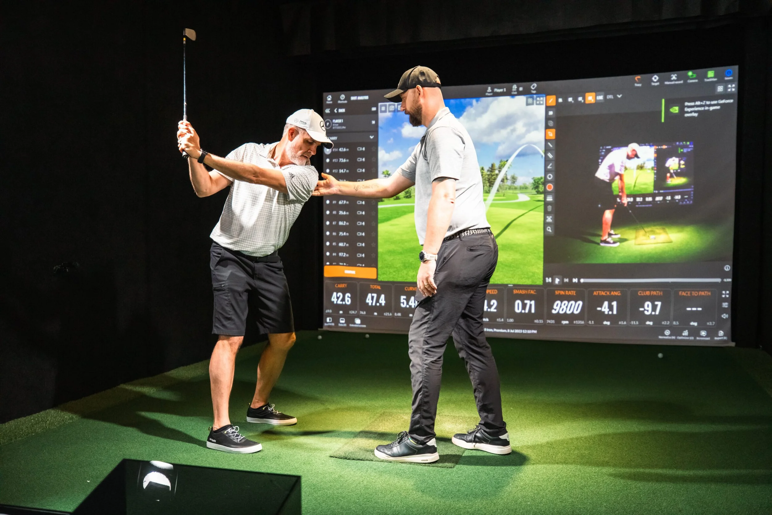 An older man taking a golf swing at a golf simulator, coached by a younger man standing beside him. The simulator screen displays a virtual golf course and various data points related to the shot.