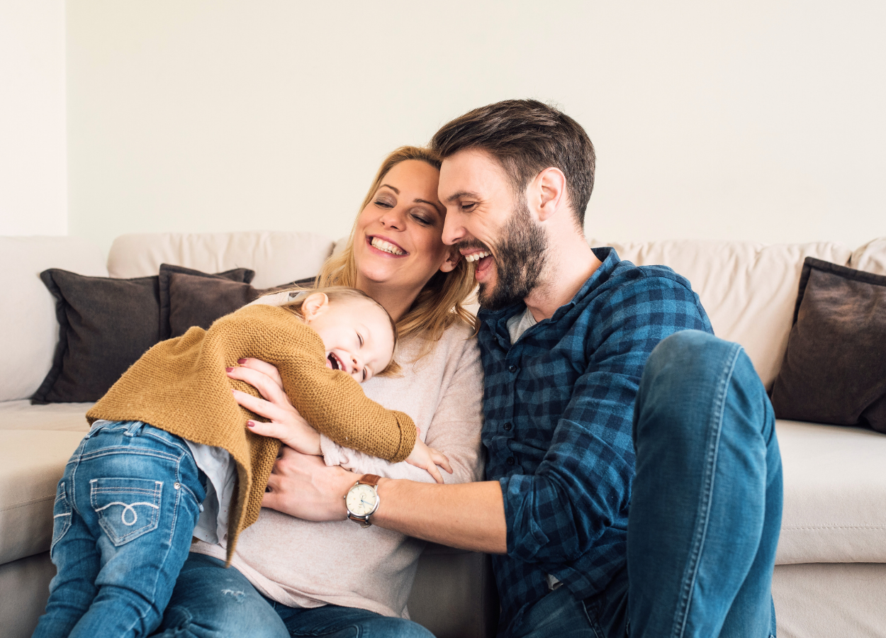 Family of three smiling and playing together on a beige couch in a living room.