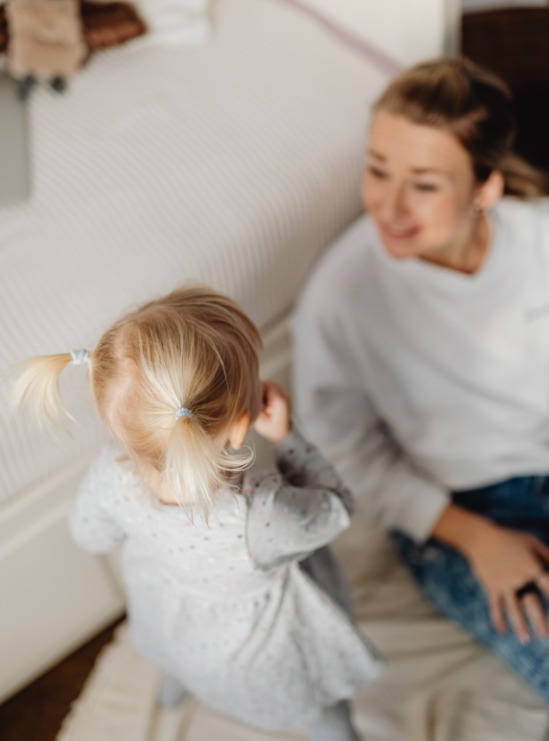 A young girl with blonde hair in pigtails talking to a smiling woman, possibly her mother, sitting on the floor in a cozy room.