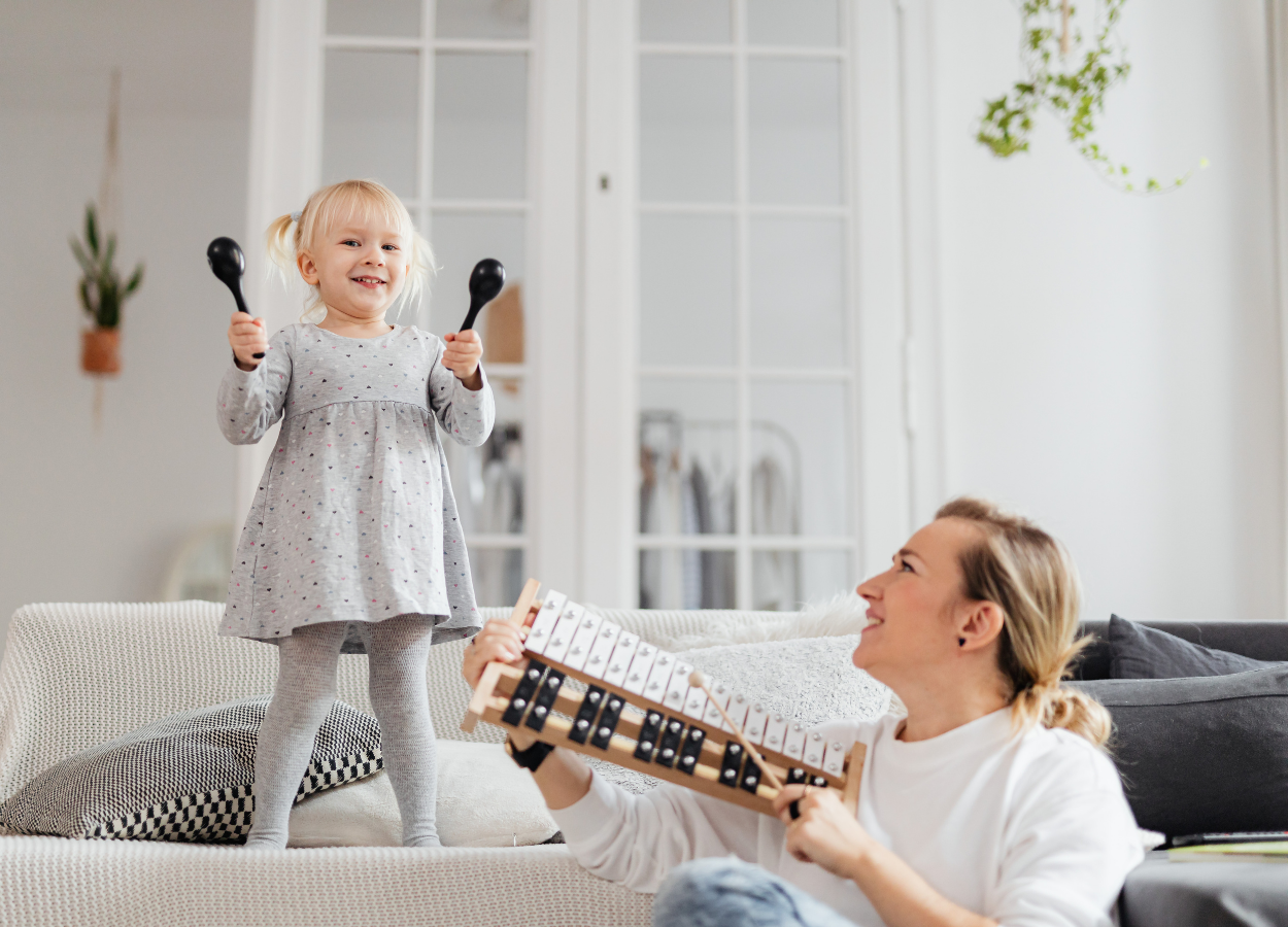 A young girl standing on a sofa holding maracas, smiling, while a woman sitting on the floor holding a xylophone looks up at her and smiles in a bright, modern living room with white walls and hanging plants.