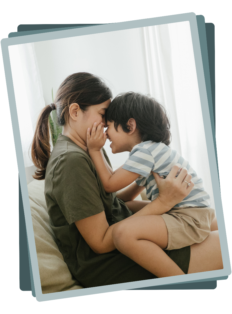 A woman and a young boy with toy face-to-face, touching noses, indoors near a window.