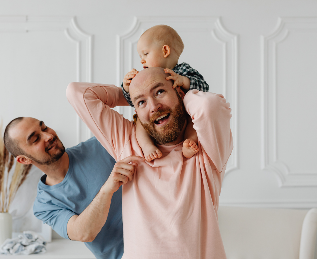 A man with a beard and pink shirt holding a young boy on his shoulders, another man standing beside them smiling in a living room.