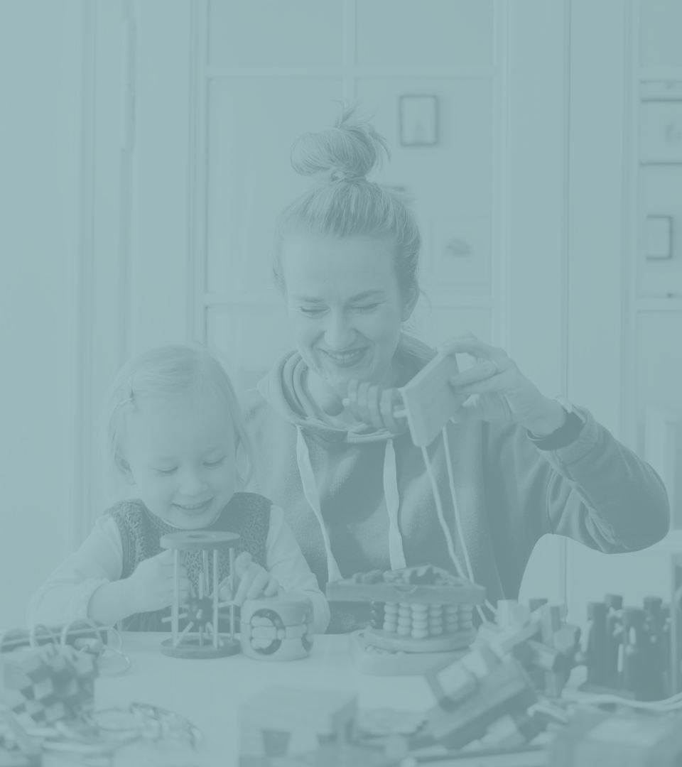 A woman and a young girl sitting at a table playing with toys and building blocks, smiling and engaging with each other.
