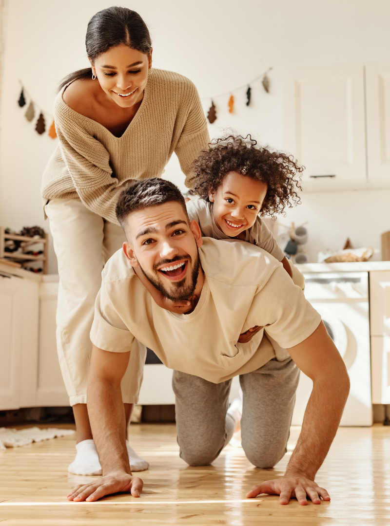 A happy family of three making a piggyback pose indoors, smiling and enjoying time together in a cozy kitchen.