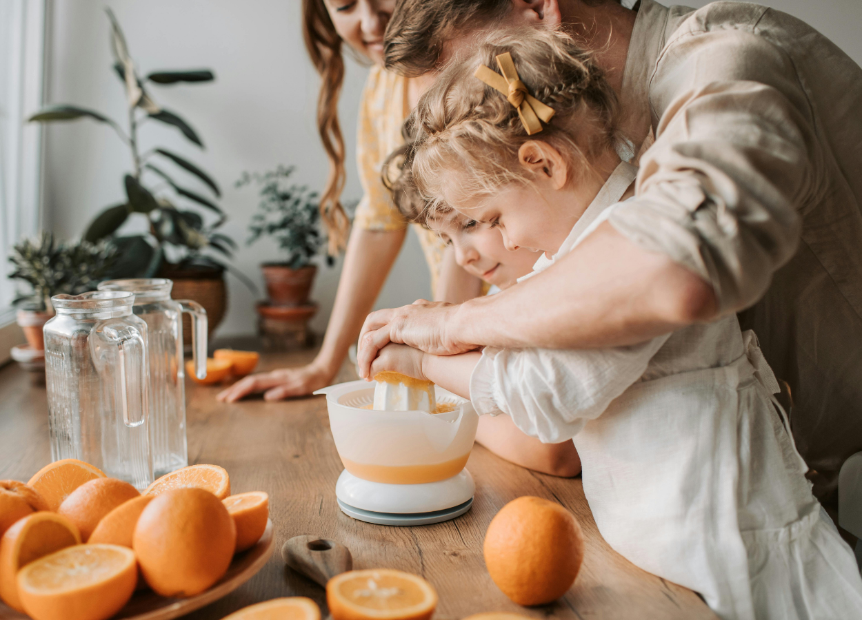 A family in the kitchen making fresh orange juice. An adult and two children are focused on squeezing oranges with a manual juicer, surrounded by whole oranges, cutting boards, and kitchen plants.