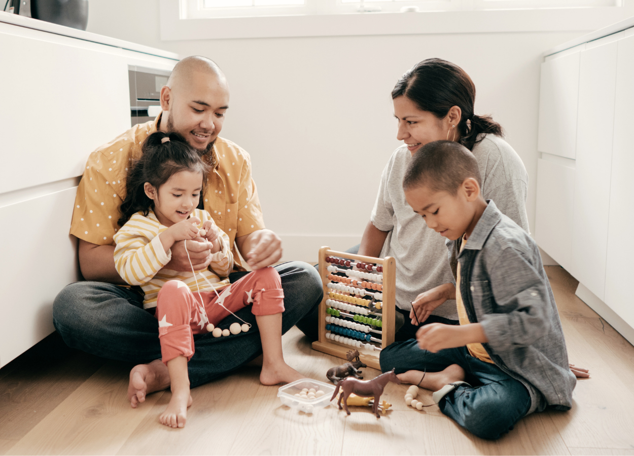 A family of four sitting on the floor playing with toys and an abacus in a bright, modern kitchen.