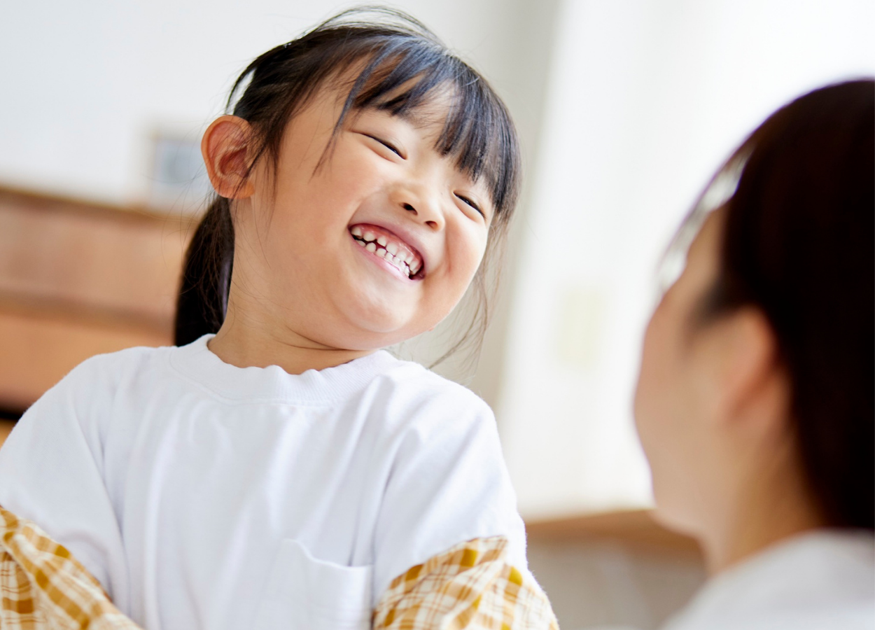 A young girl with dark hair smiling and showing her teeth, looking at a woman.