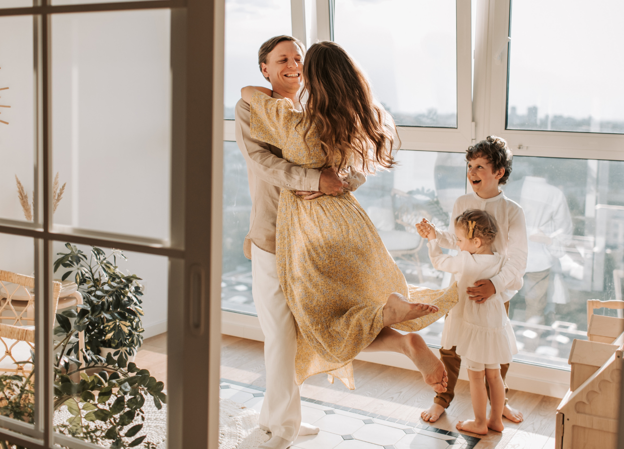 A family of four joyfully celebrating indoors, with a man lifting a woman while two children watch happily near large windows letting in natural light.