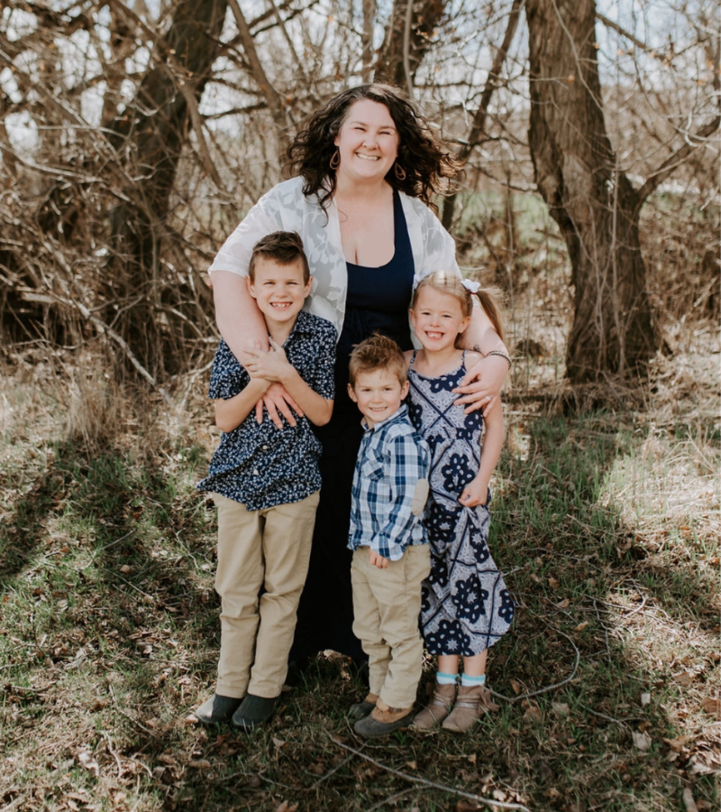 A woman with curly hair smiling and hugging three children outdoors in a wooded area, all dressed in casual clothes.