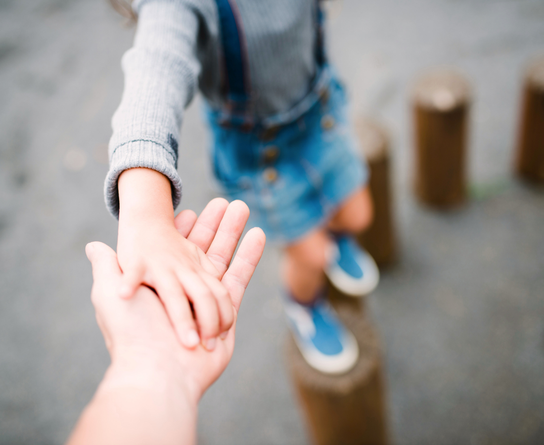 A child in denim shorts and sneakers balancing on wooden posts while holding hands with an adult.