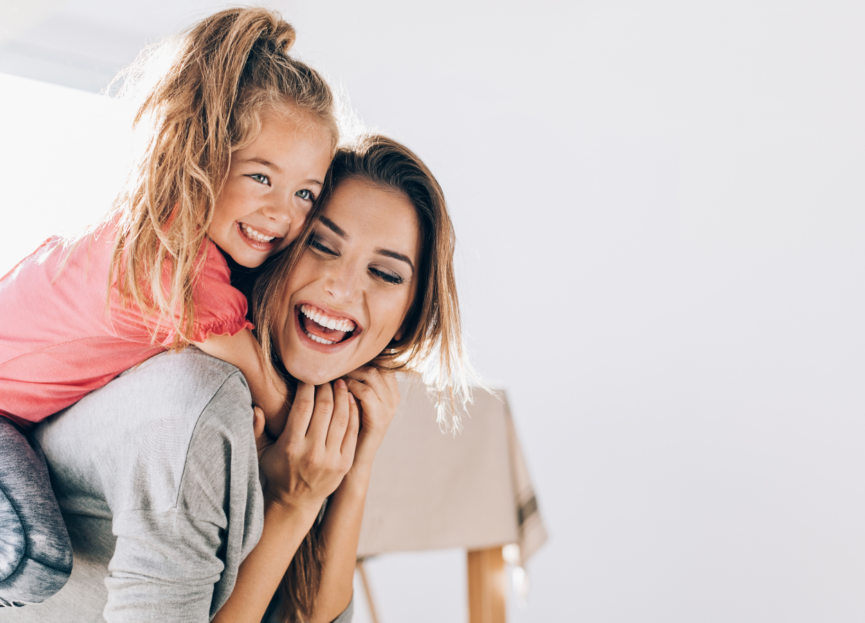 A woman with shoulder-length brown hair giving a piggyback ride to a young girl with blonde hair in a ponytail, both smiling and laughing indoors.