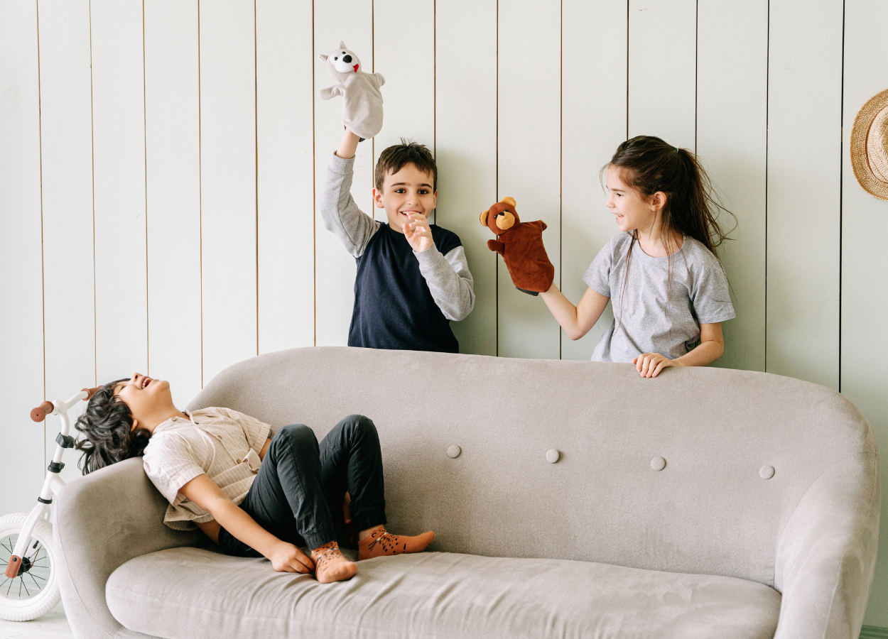 Three children playing with puppets in a living room with a light-colored sofa, a wooden wall, and a bicycle nearby. One child is lying on the sofa, laughing, while the other two are standing and holding puppets, engaging in a puppet show.