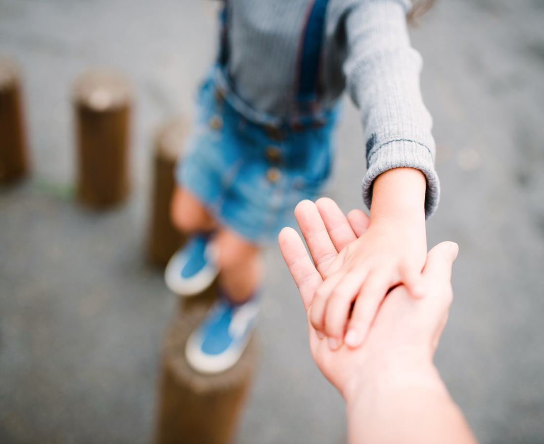 A child in a gray sweater and denim shorts balancing on a wooden post while holding hands with an adult.