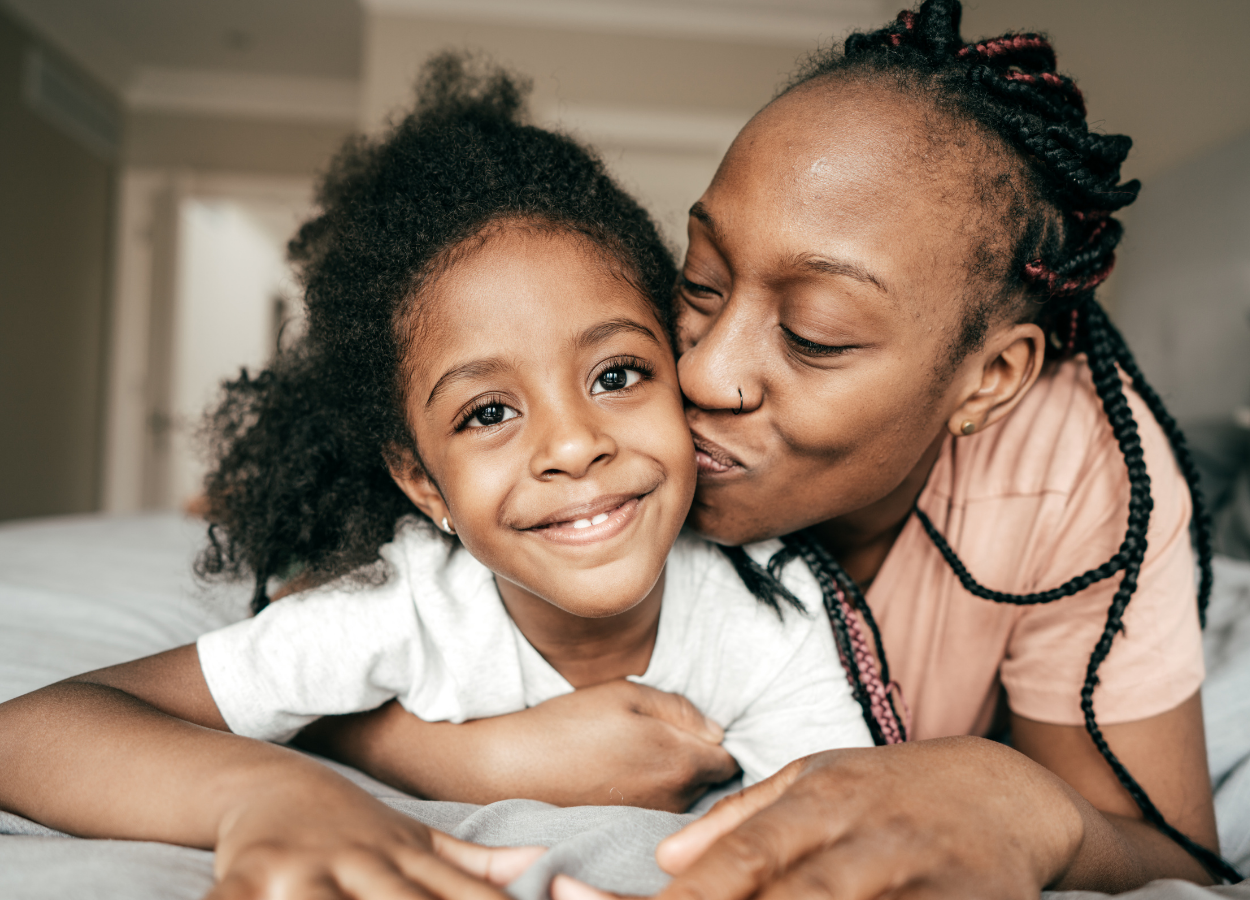 A woman with braided hair kisses a smiling young girl with curly hair on the cheek in a cozy indoor setting.