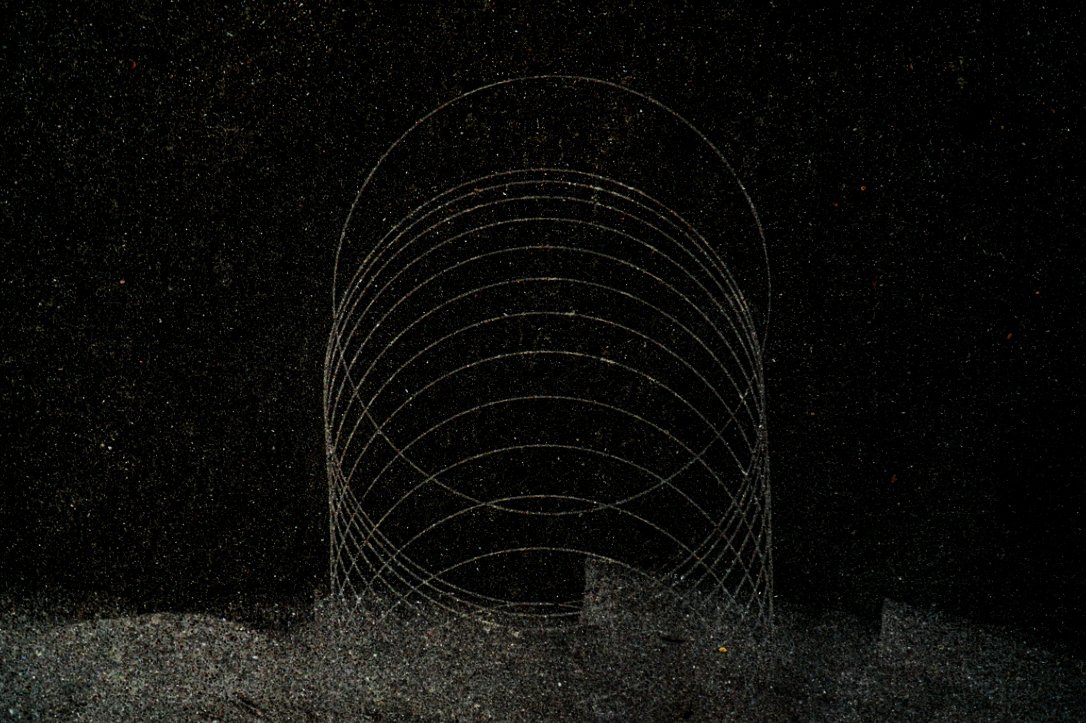 A metal wireframe sculpture of a cylinder with multiple concentric circles, set against a black background with a textured ground.