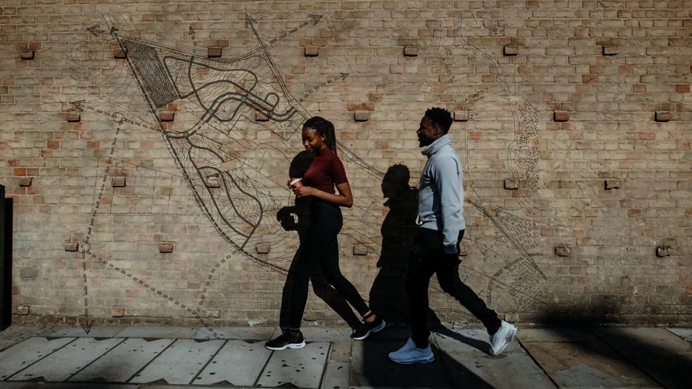 Three people walking in front of a brick wall with technical drawings and maps sketched on it, casting shadows on the wall.