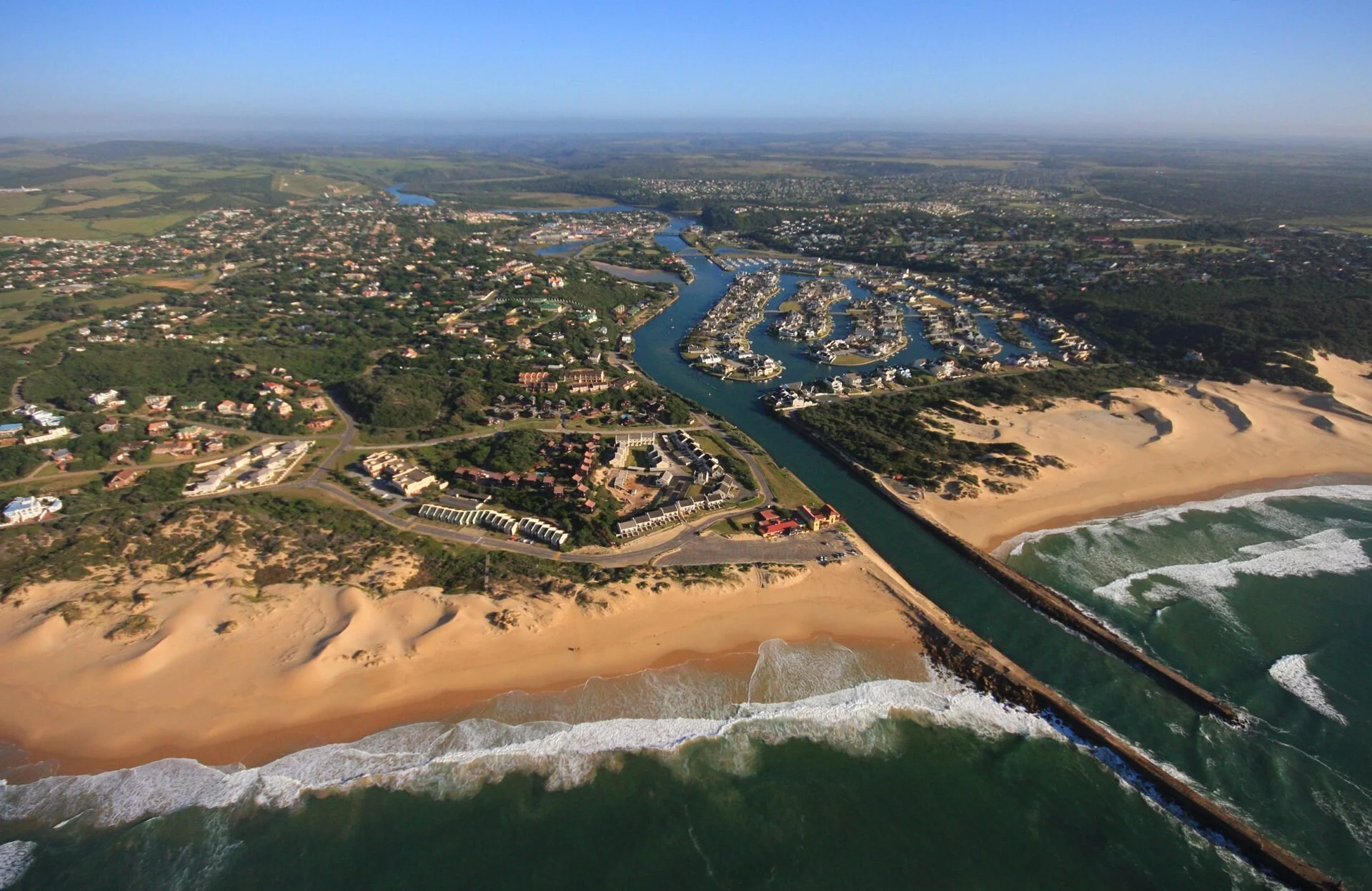 Aerial view of a coastal area with sandy beaches, a river running into the ocean, and a marina with boats. Residential buildings and greenery are visible inland.