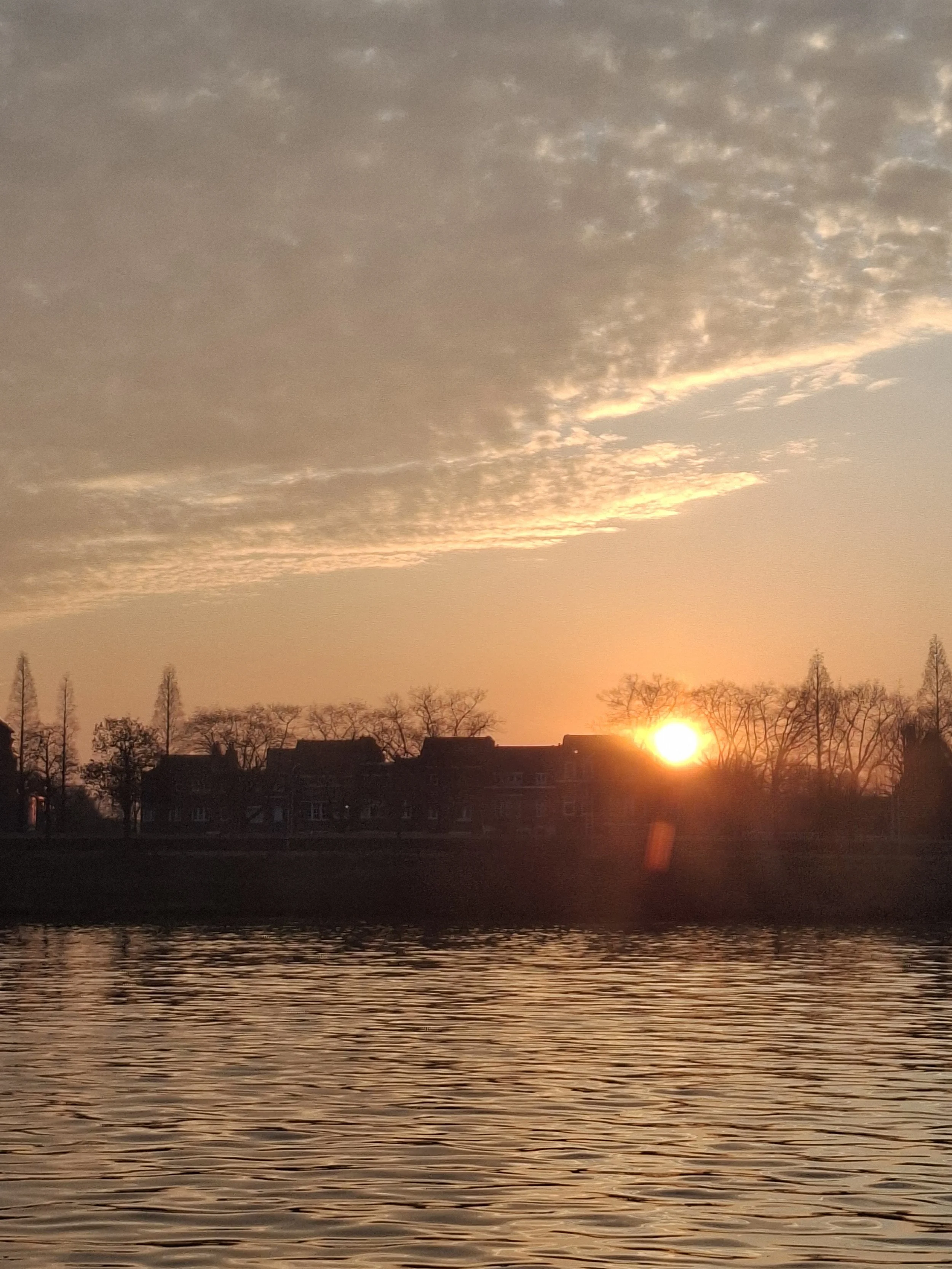 A sunset over a river with silhouettes of trees and buildings on the horizon.