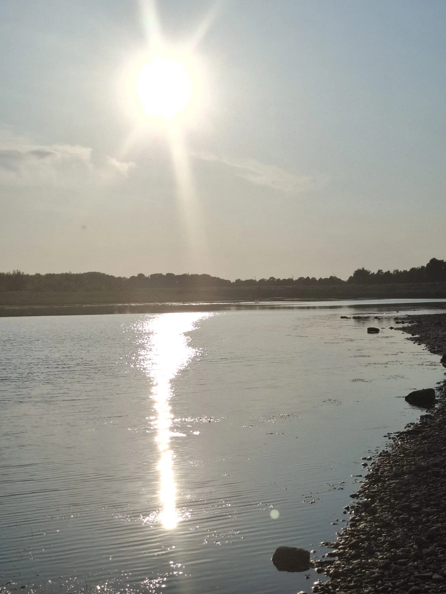 Sun shining brightly over a calm river with reflection of the sun on the water, a distant tree line, and a rocky shoreline in the foreground.