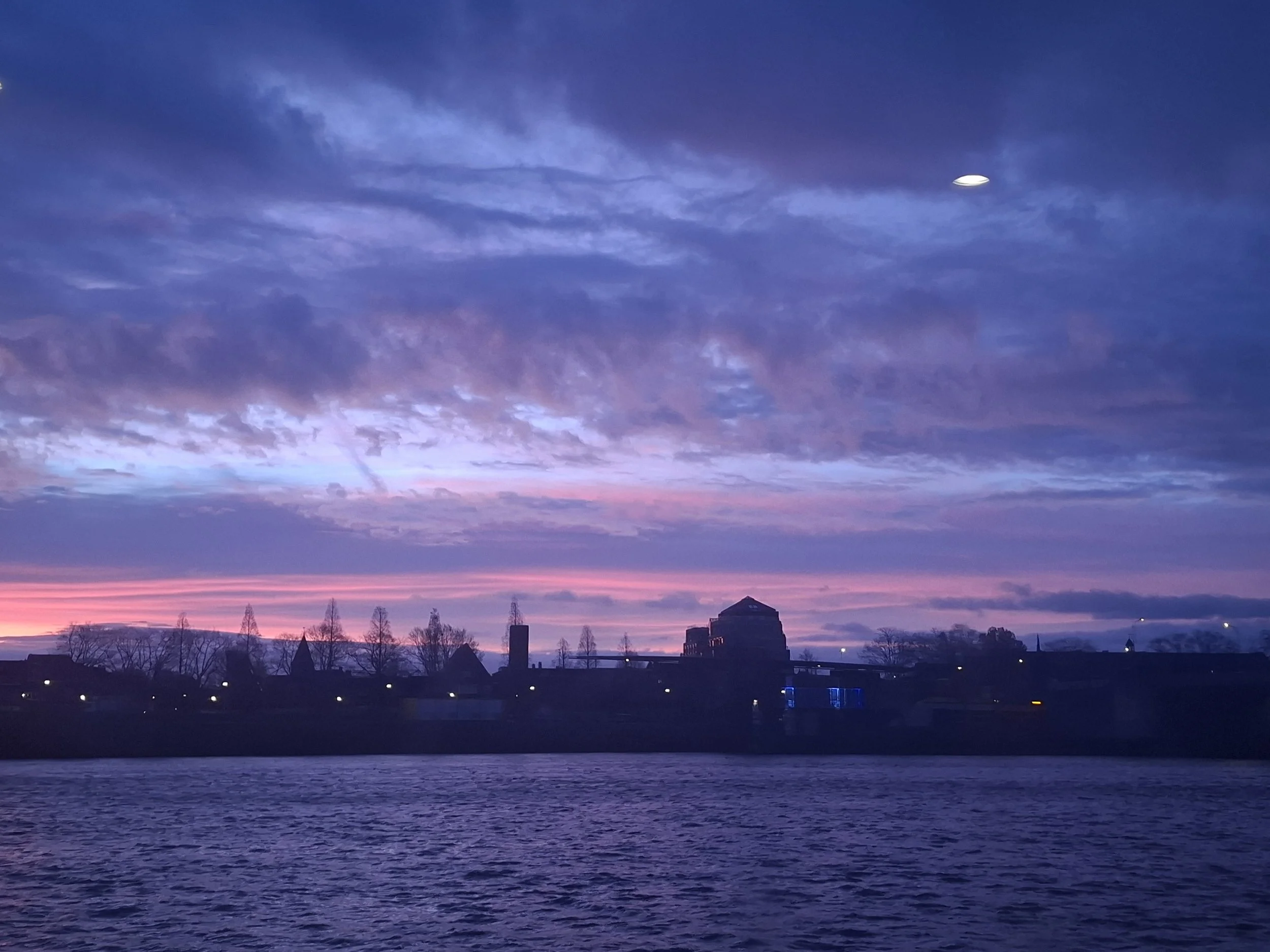 A scenic view of a city skyline at sunset with purple and pink clouds in the sky and water in the foreground.