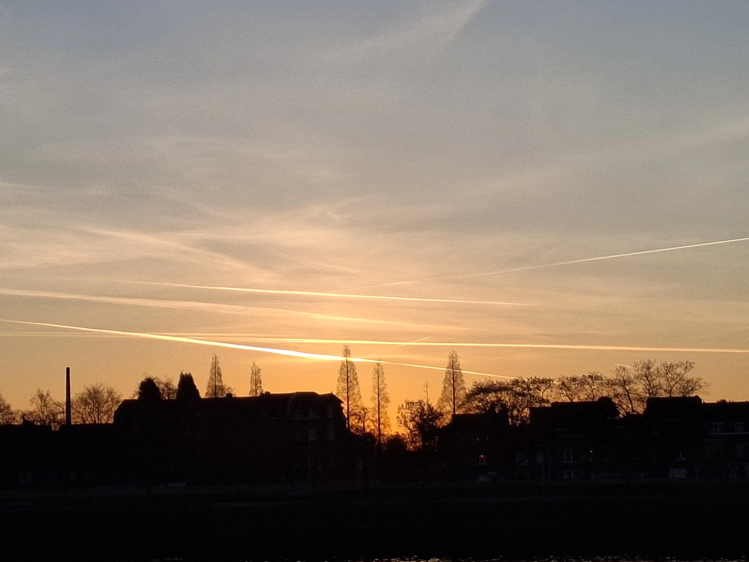 A sunset over a residential neighborhood with silhouettes of houses and trees in the foreground; the sky displays hues of orange and yellow near the horizon, transitioning to grayish tones at the top.