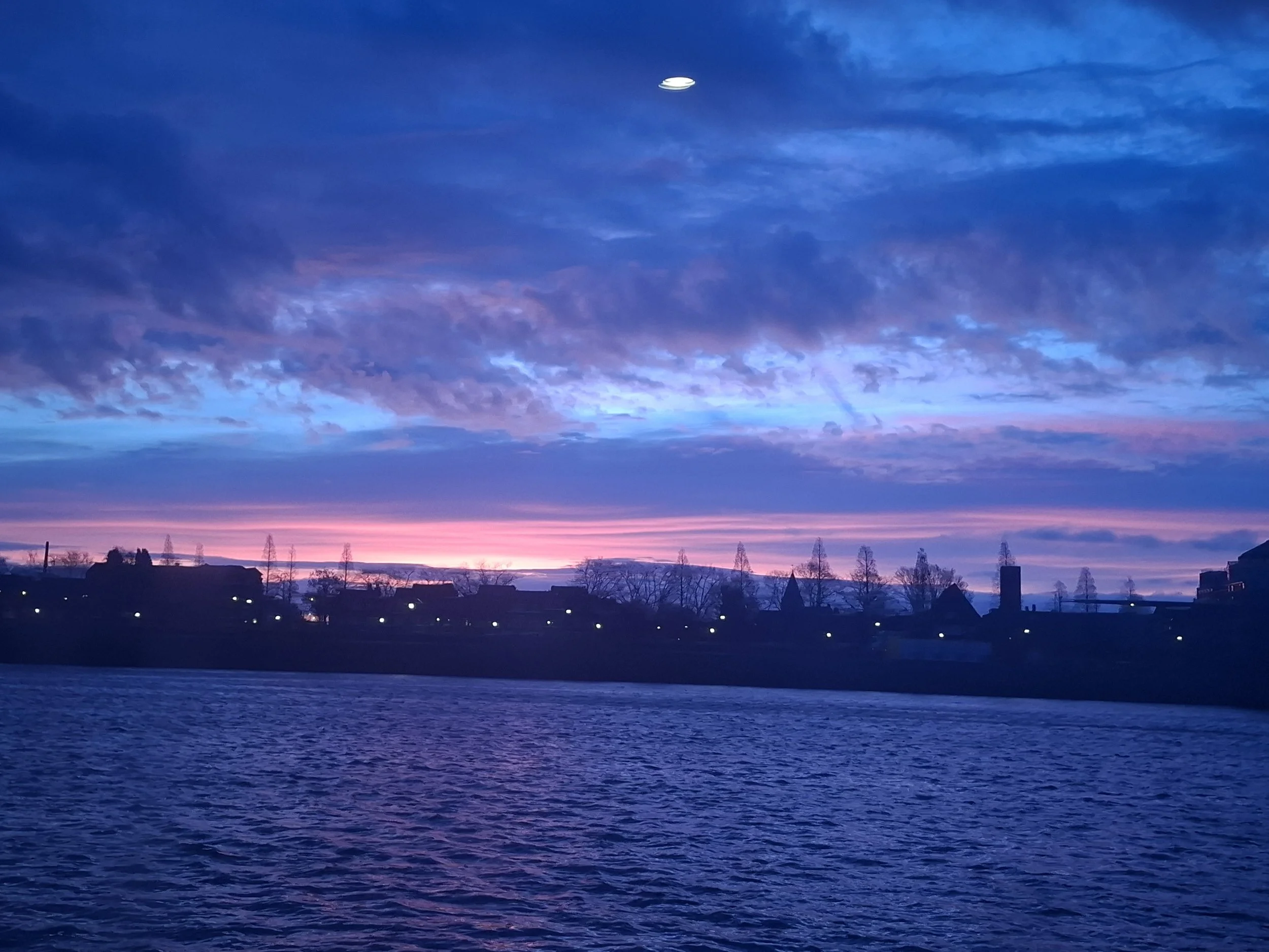 A view of a colorful sky at sunset with orange, pink, purple, and blue hues over a body of water and silhouetted trees and buildings in the distance.