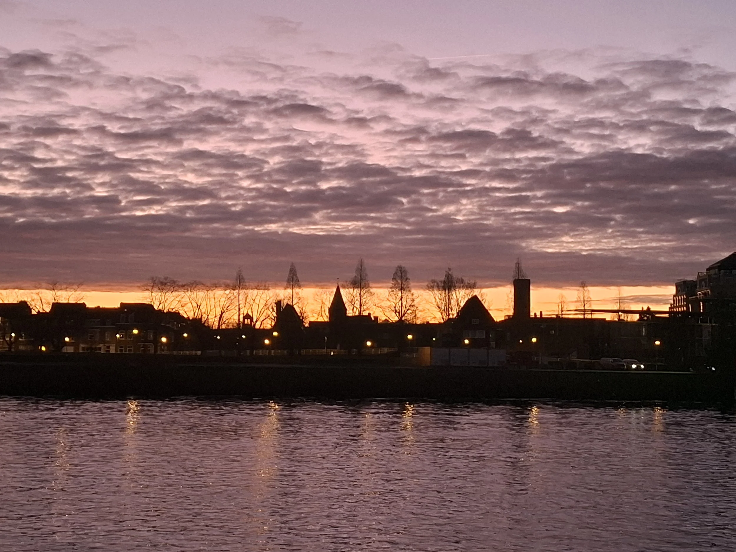 Sunset over a city skyline with a body of water in the foreground and a sky filled with pink and purple clouds.