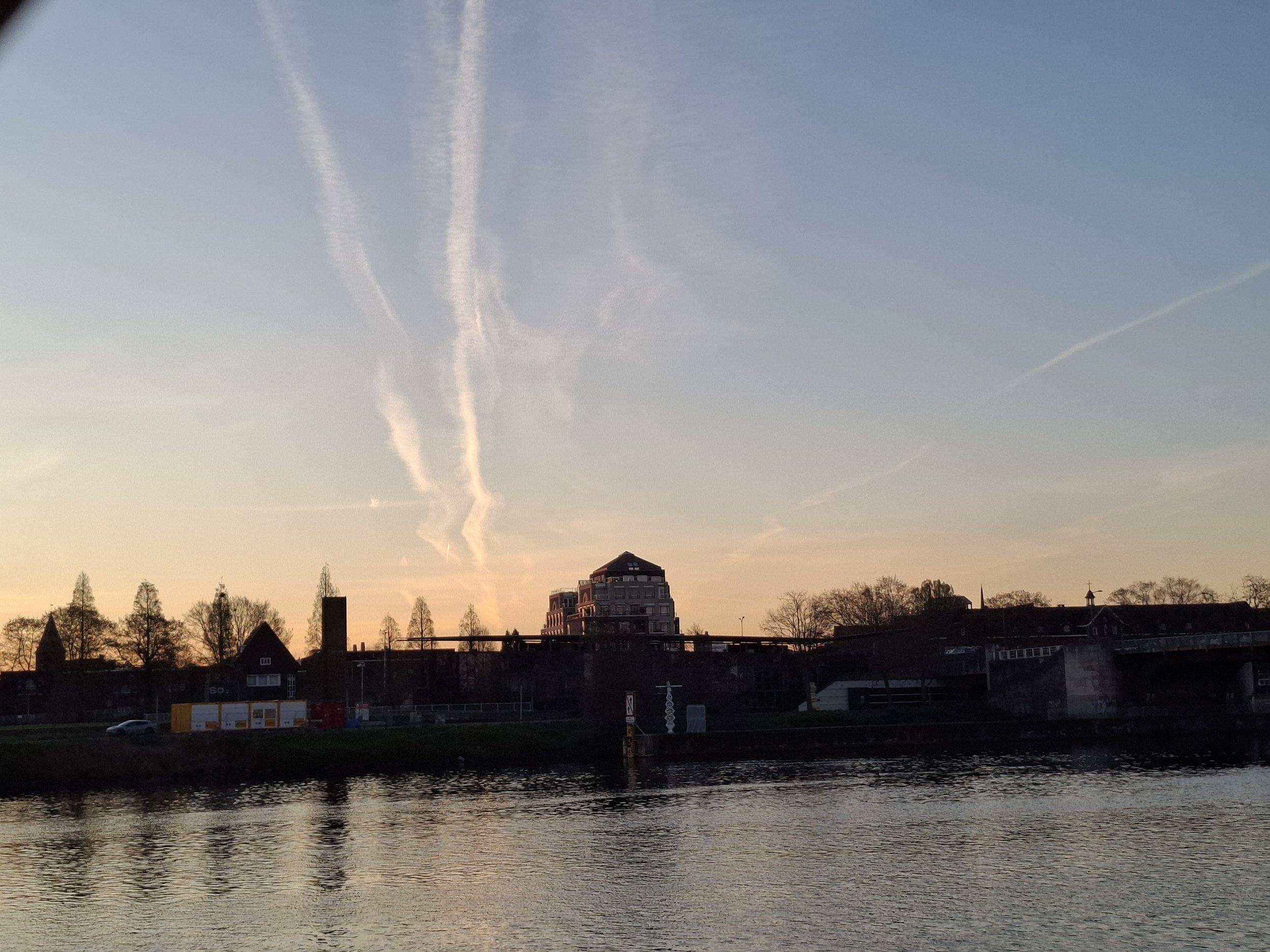 A cityscape at sunset with a river in the foreground, buildings and trees in the background, and contrails in the sky.